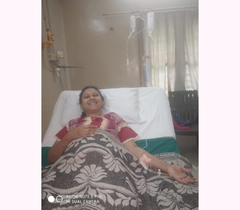 Archana Chauhan, resting in her hospital bed during her battle with cancer, wears regular clothing along with a patient mask. Her peaceful posture reflects both exhaustion and resilience, embodying the quiet strength needed to overcome adversity.