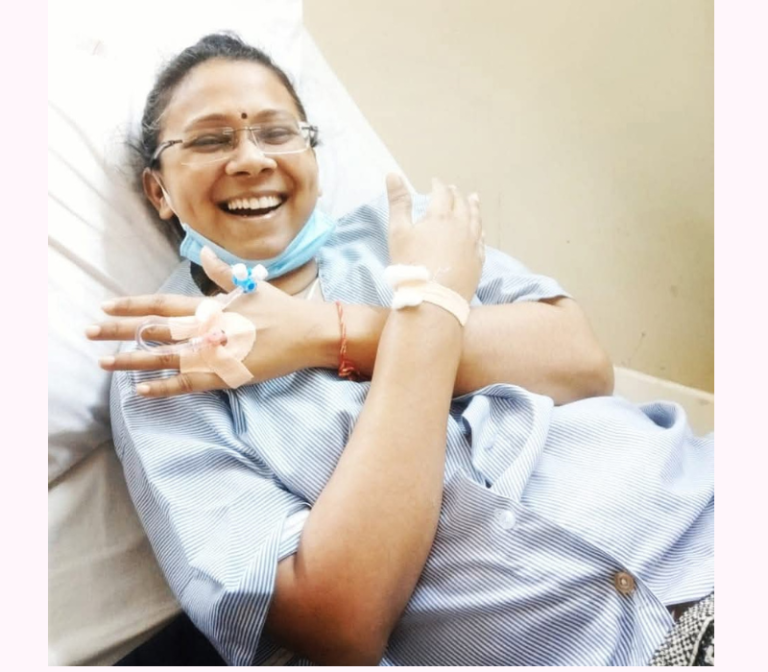 Archana Chauhan, a woman battling cancer, smiles warmly while resting on a hospital bed. She wears a blue peasant-style apron, radiating strength and positivity despite her circumstances.